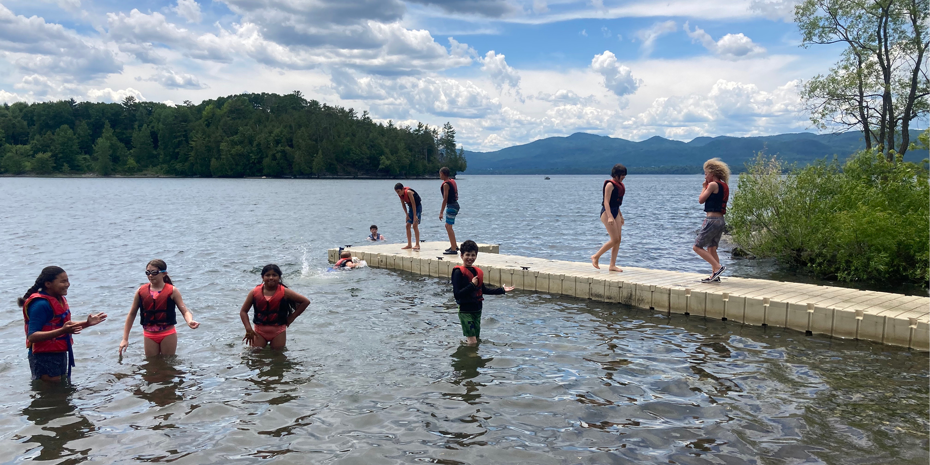 Kids playing in the lake with four kids standing knee-deep in water in the front of the photo and others visible running or swimming in the water or on a dock