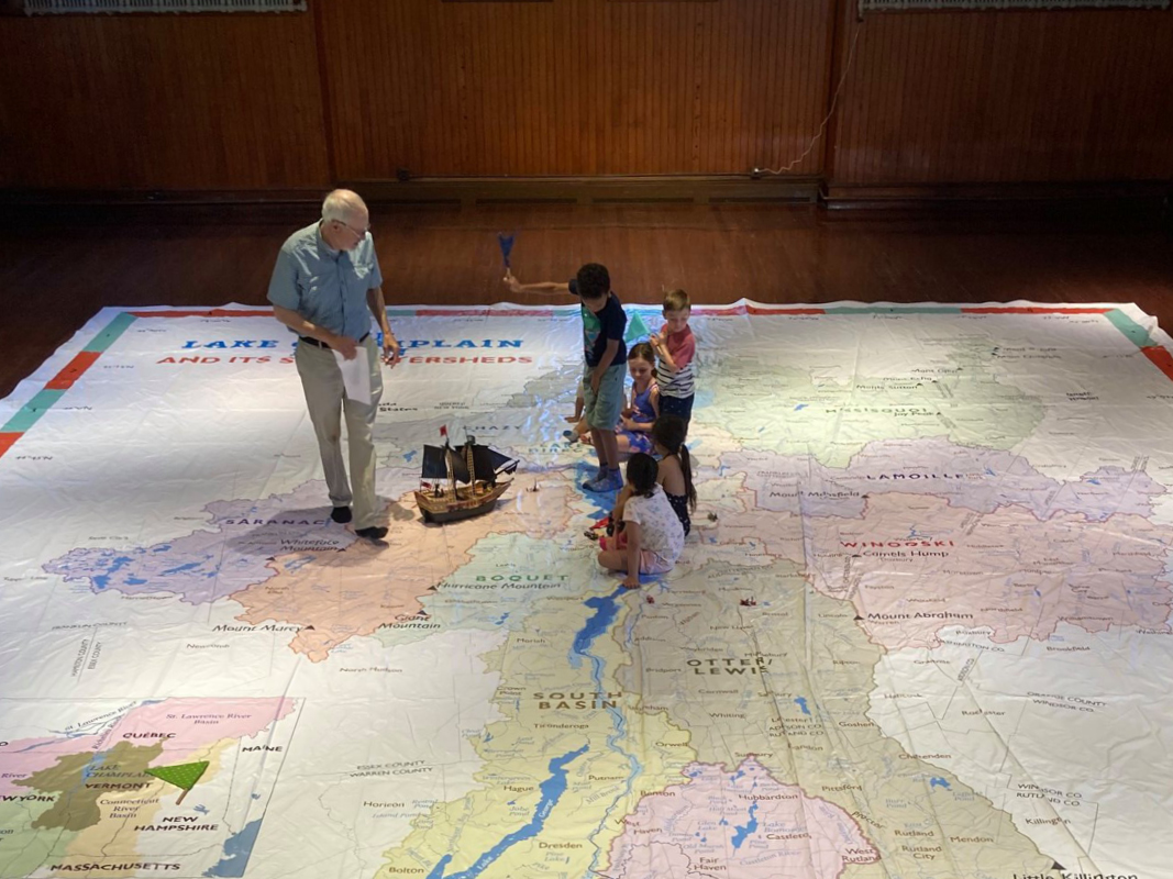 A man and four kids stand on a giant map with a small toy boat