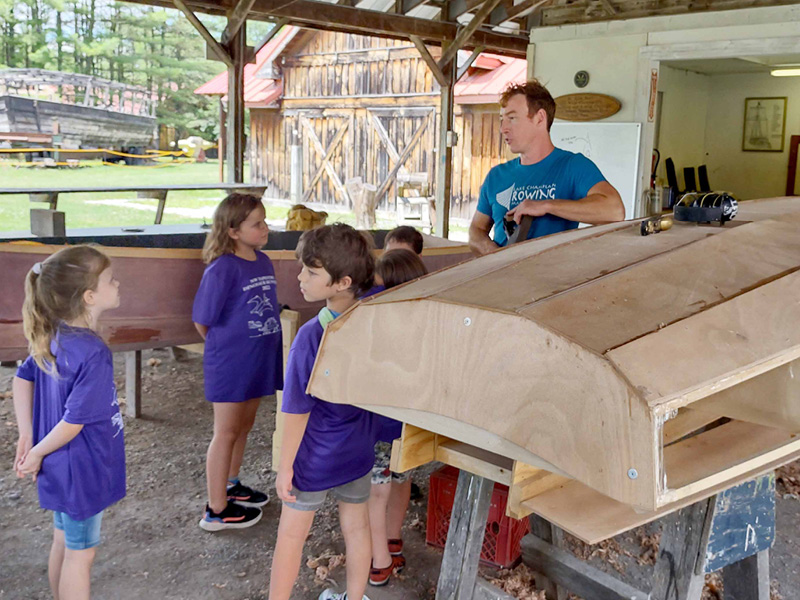 Boatbuilder Charlie Beyer talking to students about the boatbuilding process, next to several in-progress boats and materials.