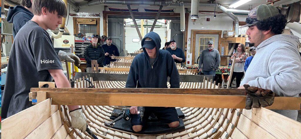 A group of students standing in and on either side of a wooden boat using tools