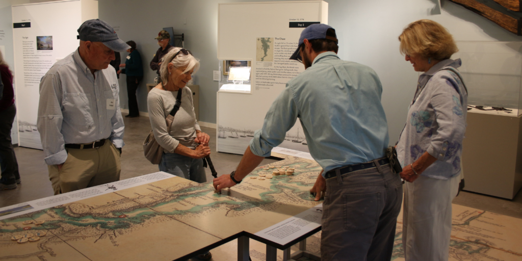 Four adults look at a large map raised on a table in an exhibit building at Lake Champlain Maritime Museum