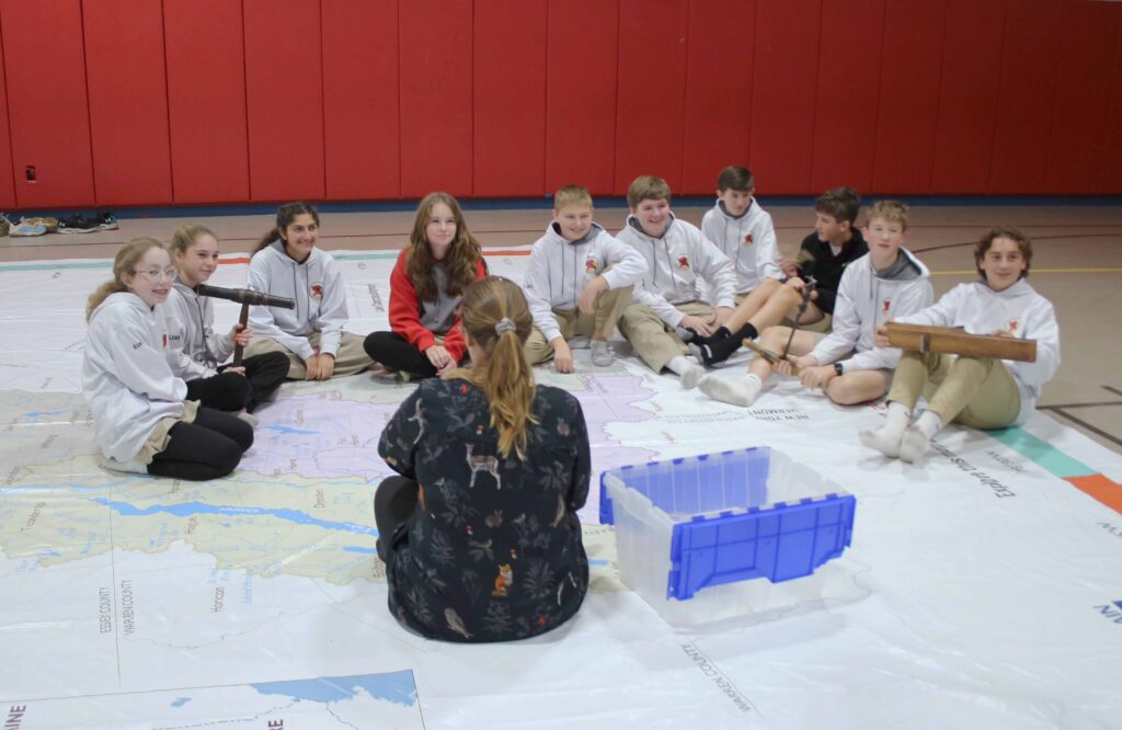 Teenage students sitting around a teacher on a large map on the floor.