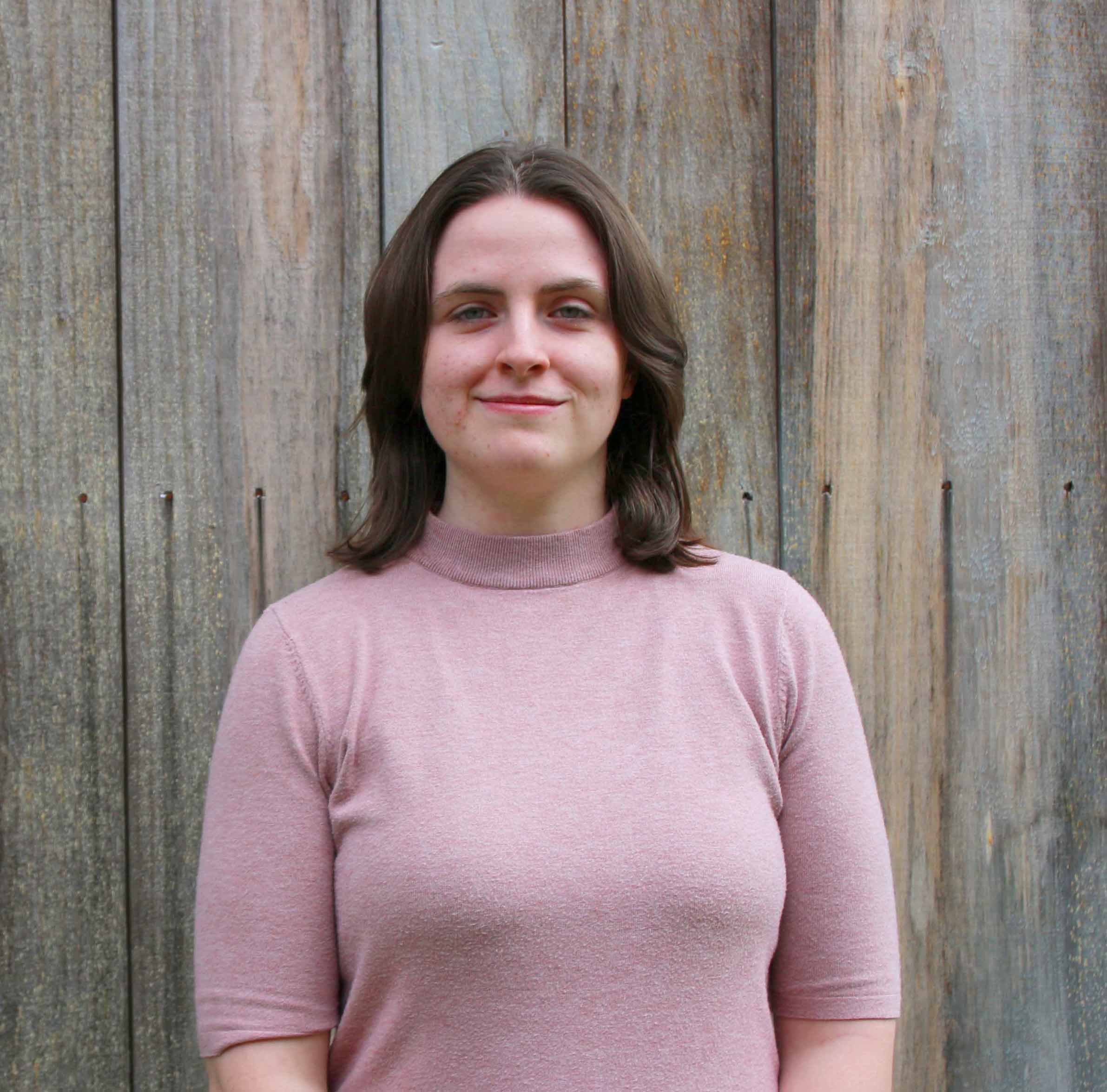 Person with shoulder length dark hair and light pink shirt in front of a wooden wall