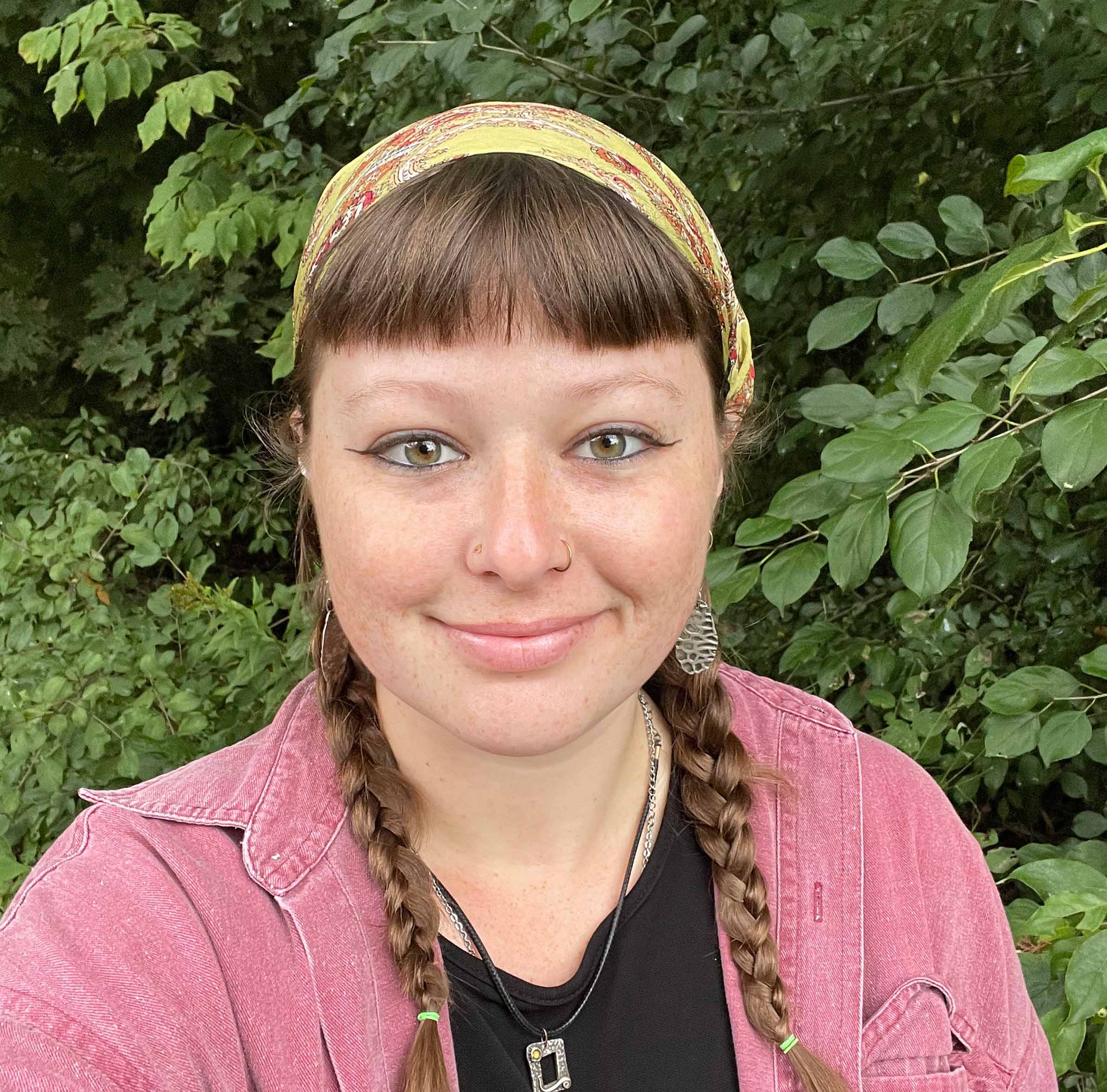 A person with two braids, yellow headband, and pink jacket in front of green leaves